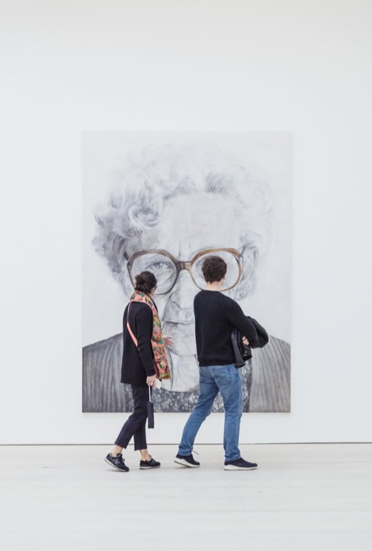 Grande retrato de idosa em preto e branco com duas pessoas olhando para o quadro